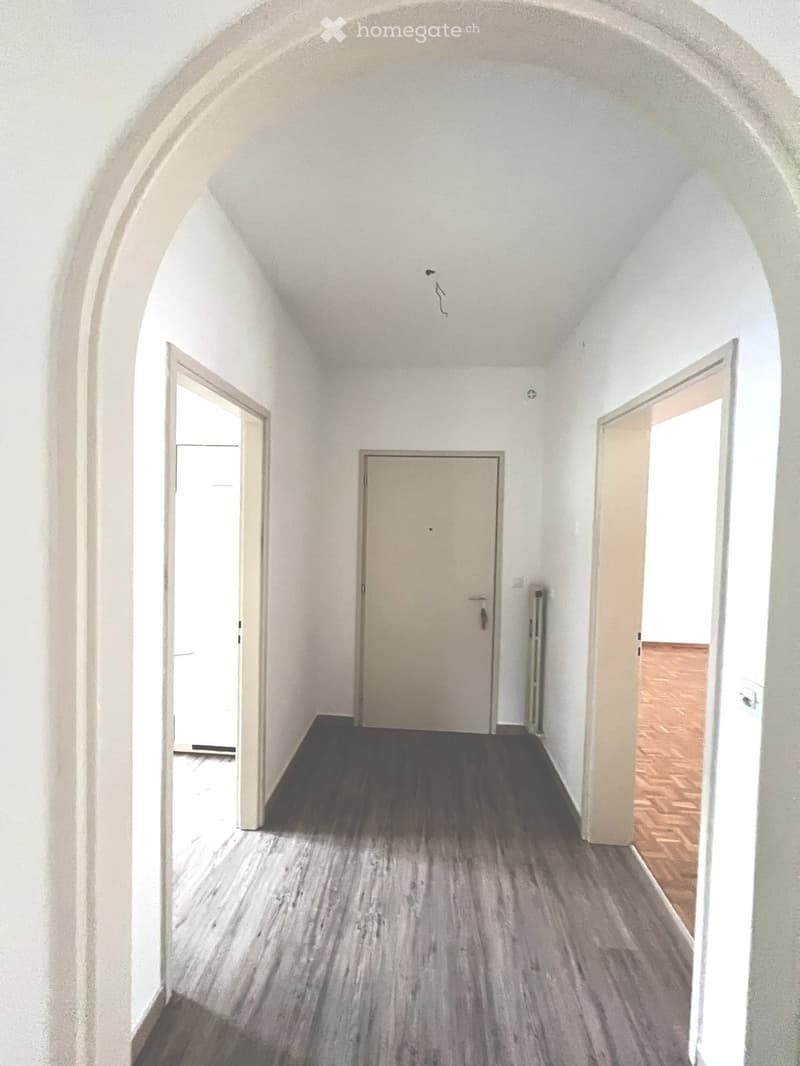 The image shows a hallway with a white arched ceiling, white walls, and a gray laminate floor. The hallway has a door at the end and another door on the right side. The hallway appears to be in a residential or apartment building.