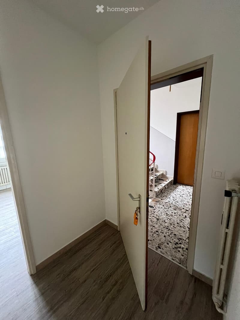 The image shows a hallway with white walls, a wooden floor, and a doorway leading to another room with a terrazzo floor.
