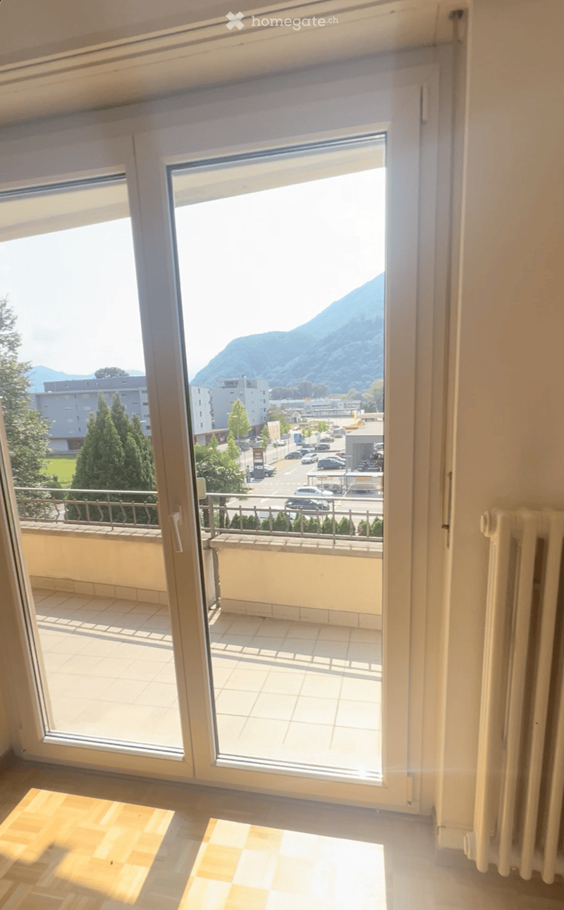 The image shows a room with a large window that provides a view of a city skyline and surrounding mountains. The room has a balcony or terrace accessible through the window. The floor appears to be tiled, and there is a radiator visible in the room.