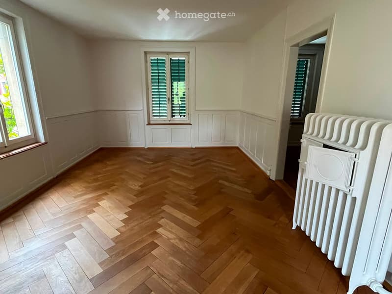 Empty room with wooden herringbone flooring, white walls, and large windows with shutters. A white radiator is visible on the right side.