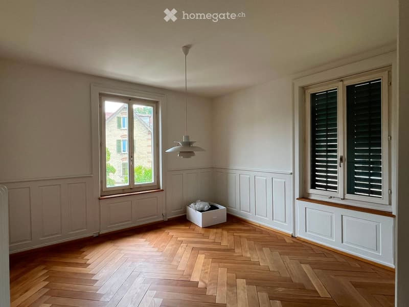Empty room with wooden herringbone flooring, white walls, and large windows with shutters. A single pendant light hangs from the ceiling.