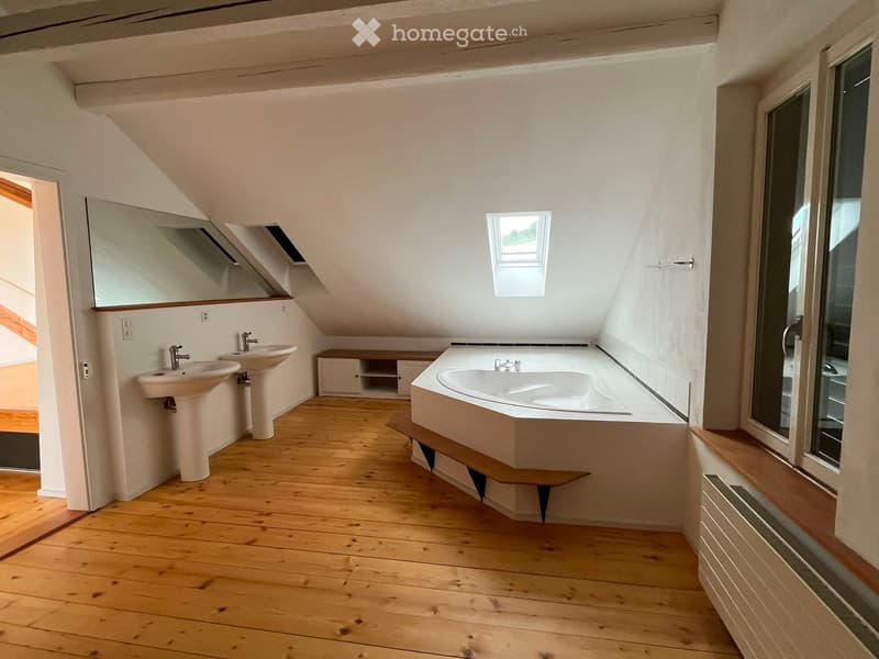 Bright, spacious bathroom with wooden flooring, two sinks, a bathtub, a large mirror, and a radiator. The room features skylights and a slanted ceiling.