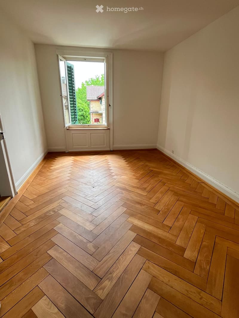Empty room with herringbone-patterned wooden flooring, light-colored walls, and a large window with a view of greenery and neighboring buildings.