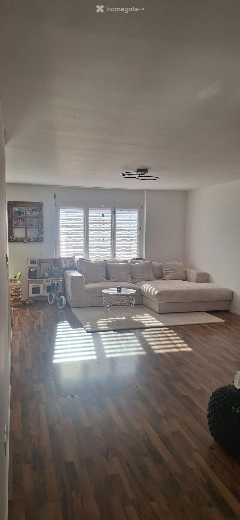 Modern living room with a large sectional sofa, coffee table, and wooden flooring. The room features large windows with blinds and a small kitchen area visible in the background.