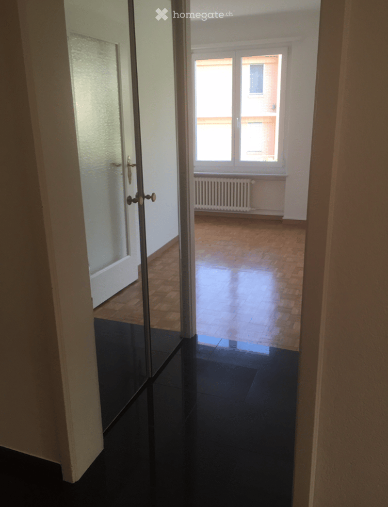 Empty room with wooden flooring, large window, radiator, and glass door leading to another room.