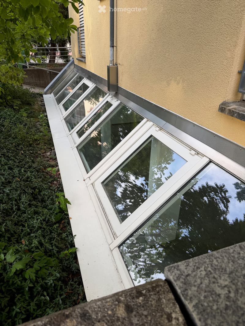 A modern glass balcony terrace with multiple large, angled glass panels, attached to a yellow building exterior, surrounded by greenery.