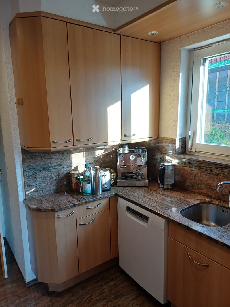 Modern kitchen with light wooden cabinets, granite countertops, and stainless steel appliances including a sink, dishwasher, coffee maker, toaster, and water kettle.