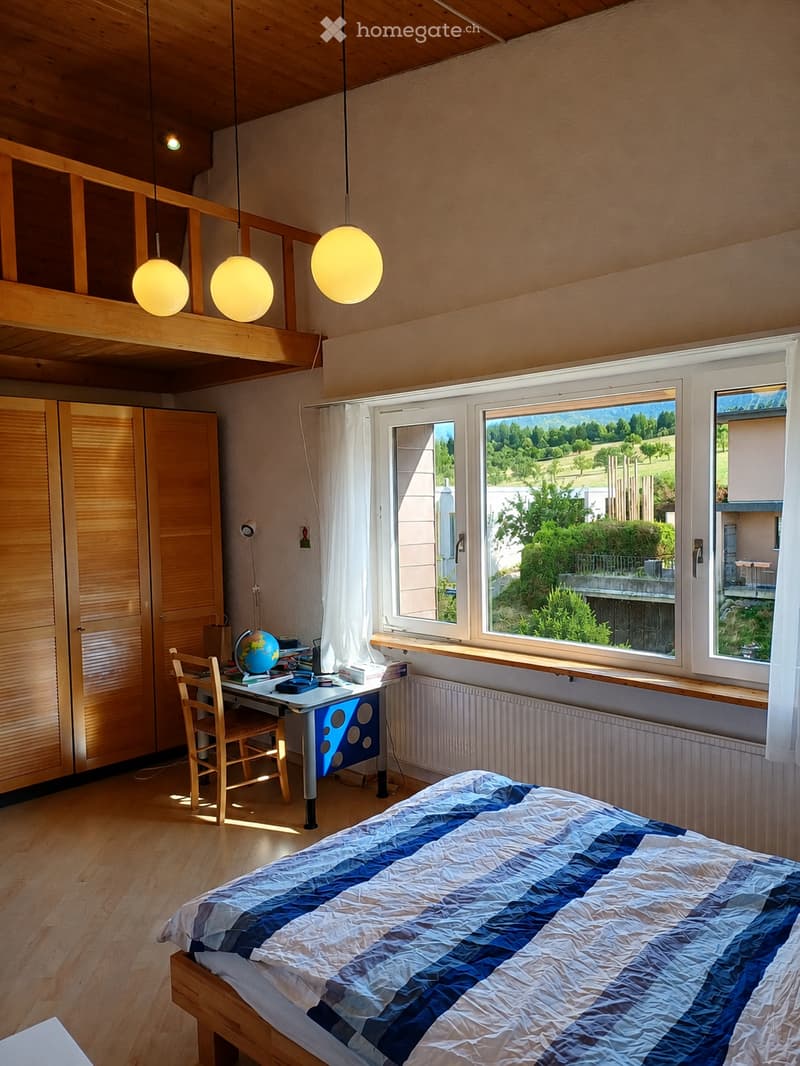 Bright bedroom with wooden floors, large windows overlooking a green landscape, a bed with a blue and white quilt, a desk with a globe, and a wooden wardrobe.