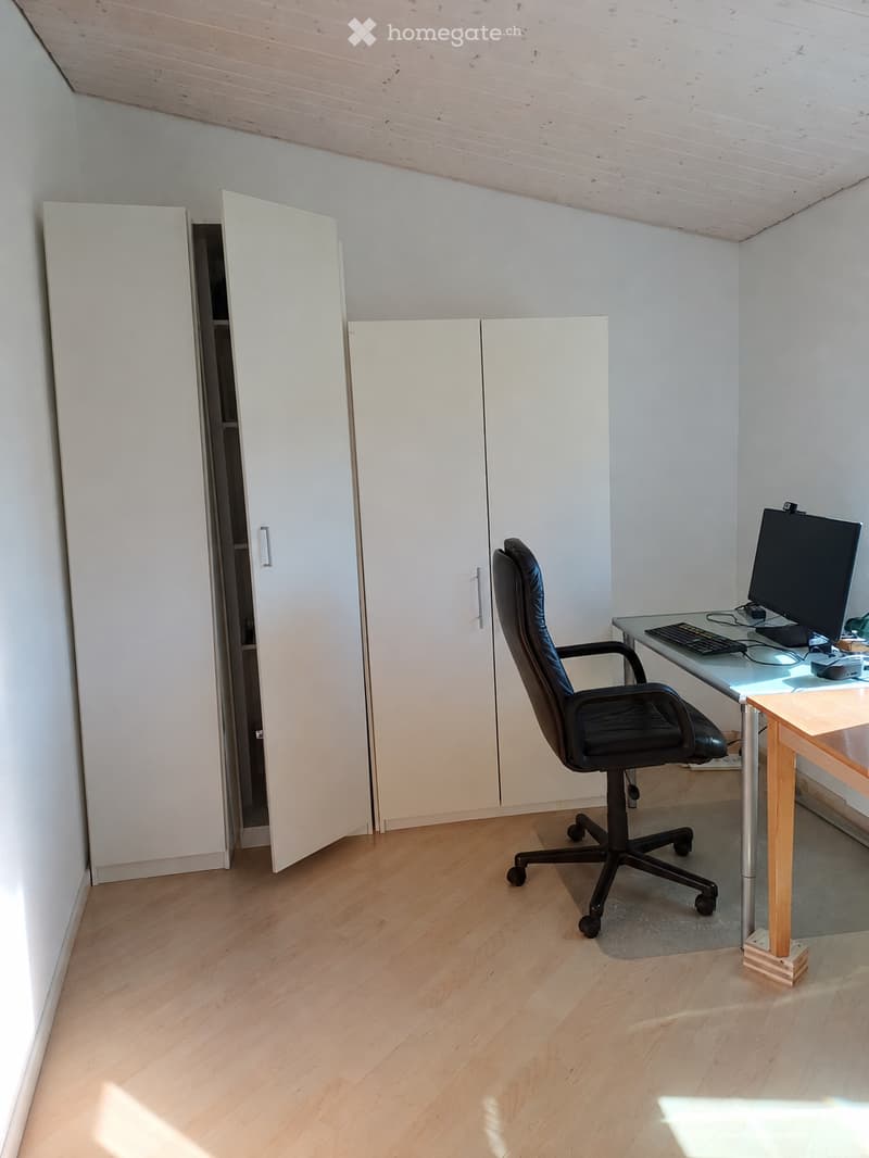 A modern, minimalist office space with light wooden flooring, white walls, and built-in white cabinets. A black office chair is positioned in front of a sleek glass desk holding a computer, keyboard, and mouse.