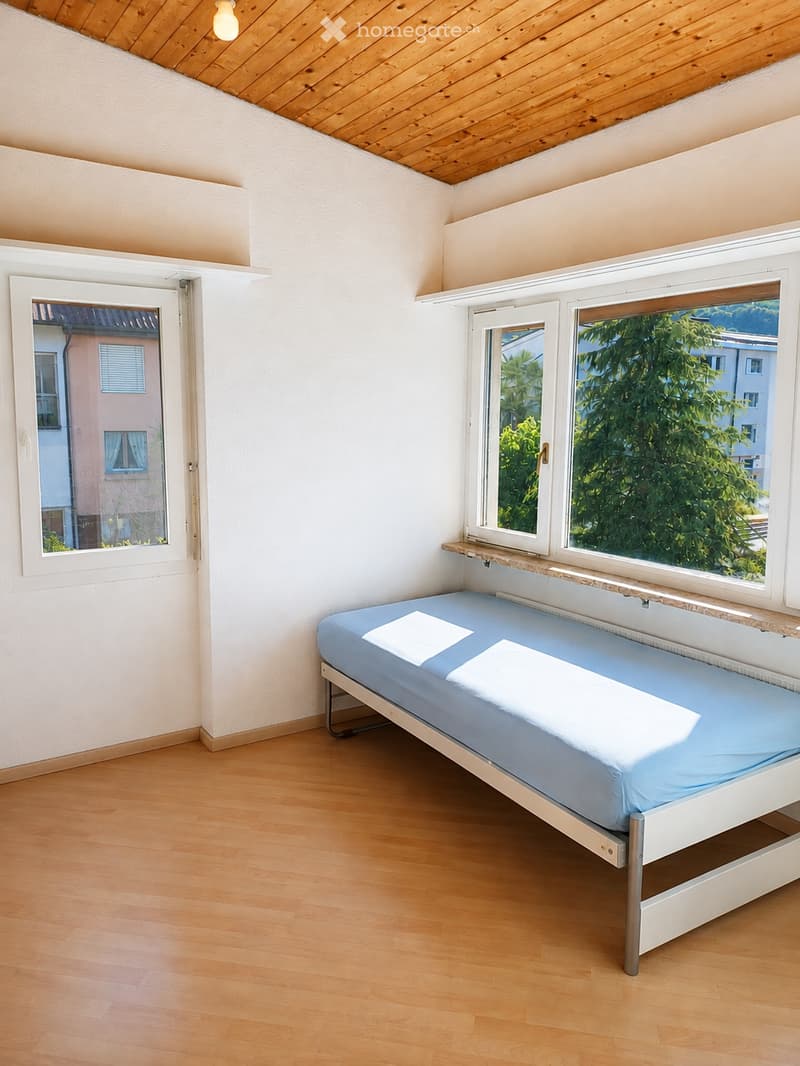 Minimalist bedroom with a single bed, light blue bedding, wooden flooring, and large windows offering a view of greenery outside.
