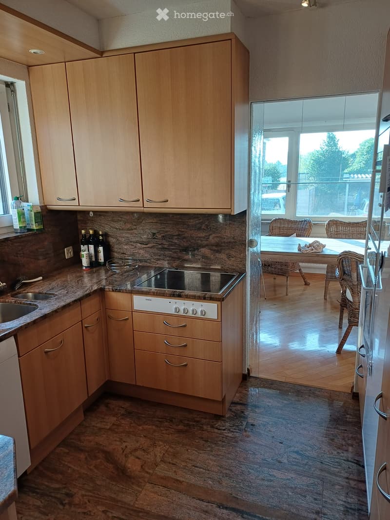 Modern kitchen with light wooden cabinets, granite countertops, and a central electric stove. A window offers a view of a dining area with wicker chairs and a table.