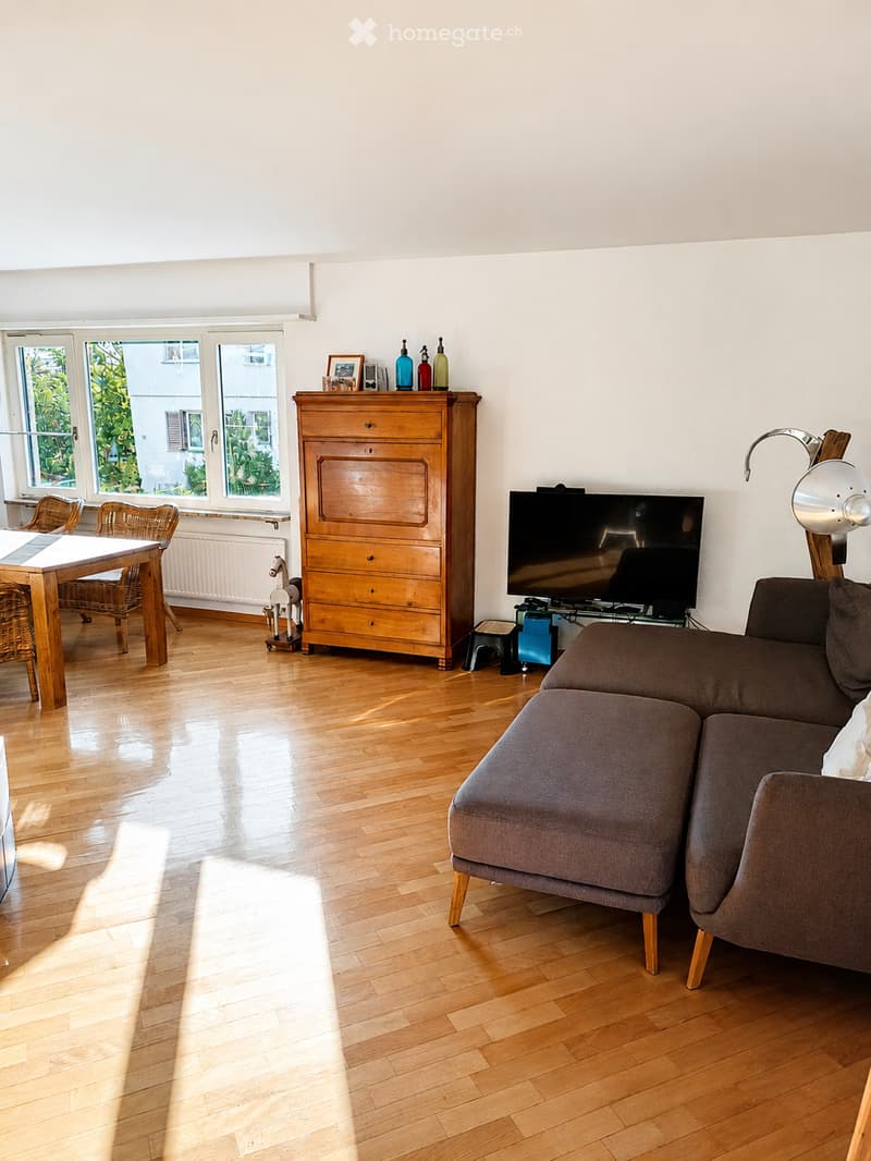 Bright living room with wooden flooring, large windows, a grey sofa, a wooden dining table with chairs, a television, a wooden cabinet, and a small toy horse.