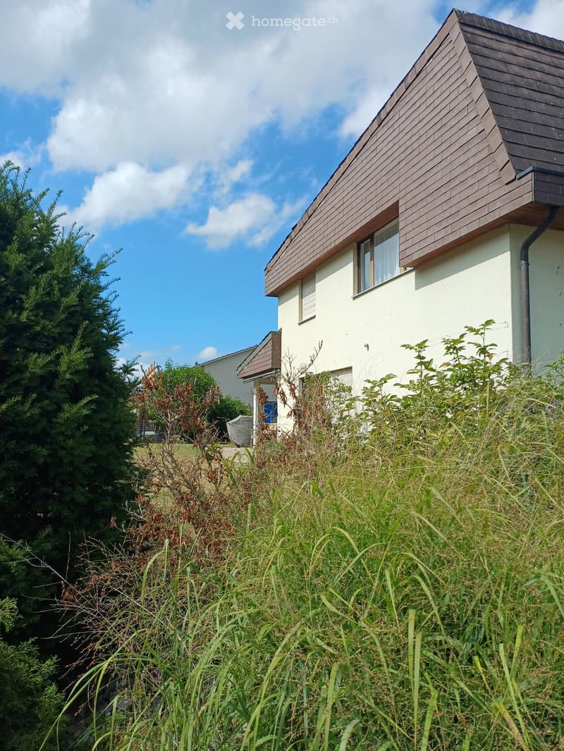 Modern two-story house with white walls and brown wooden roof, surrounded by greenery and tall grass.