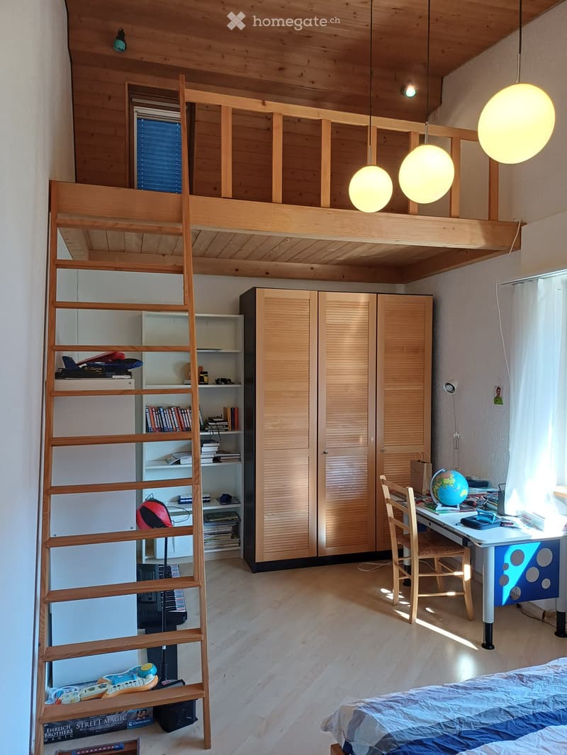 A modern, multi-functional bedroom with a lofted sleeping area, a desk, and a bookshelf. The room features wooden floors, a large window with white curtains, and several hanging pendant lights.