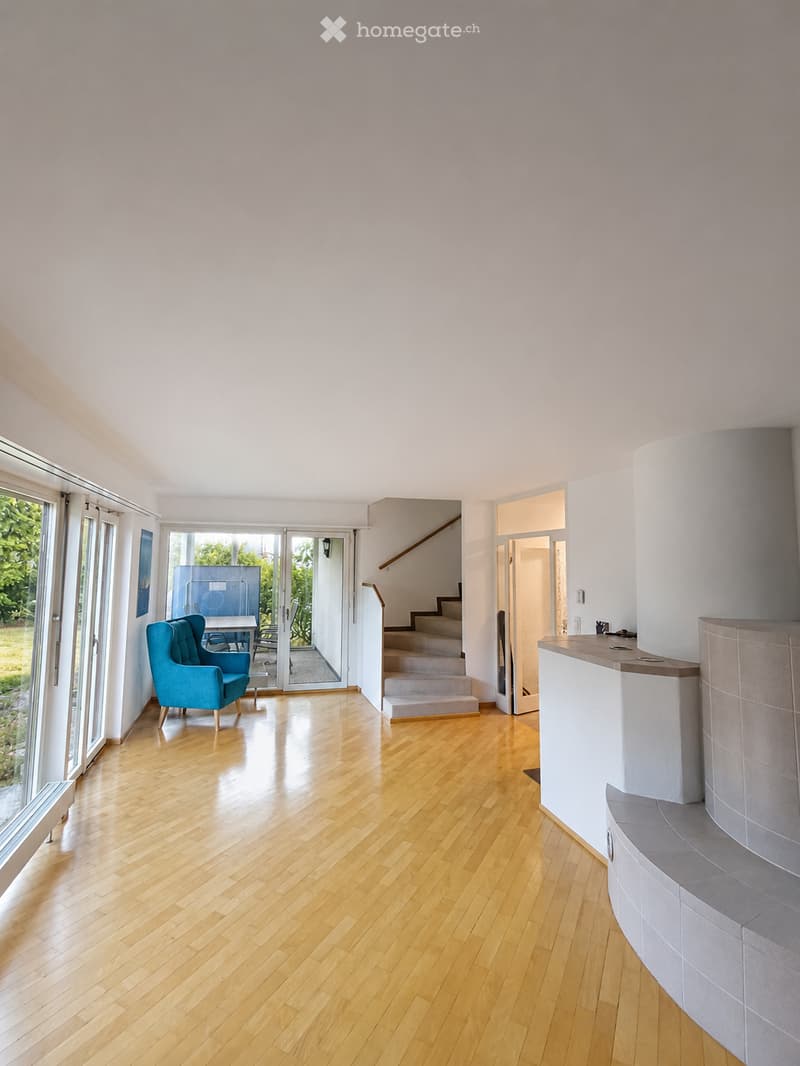 Bright, open living space with wooden flooring, large windows, a staircase leading upstairs, and a blue armchair near a small table.