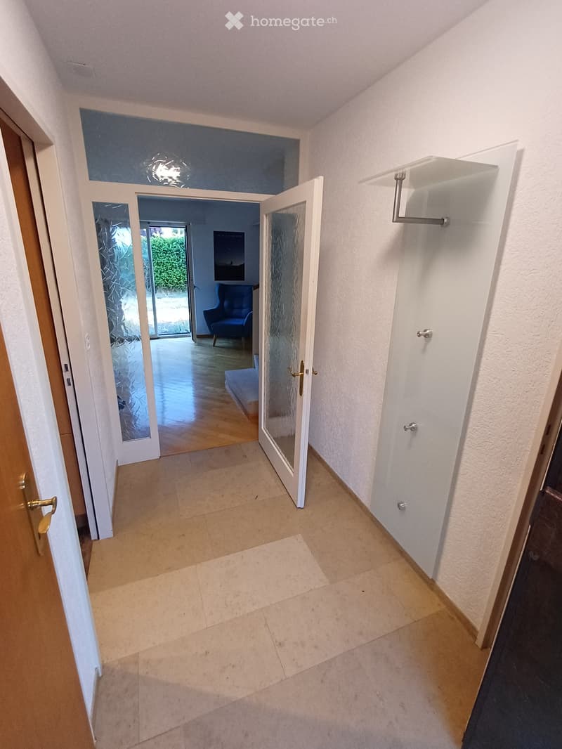 Empty hallway with tiled floor, white walls, and glass doors leading to another room with wooden flooring and a blue armchair visible in the background.