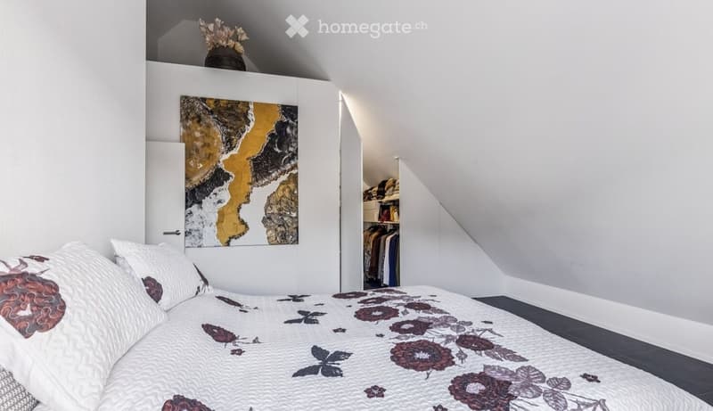 Bedroom with white walls, bed with floral comforter, white pillows, painting on wall, closet with clothes, vase with flowers on top