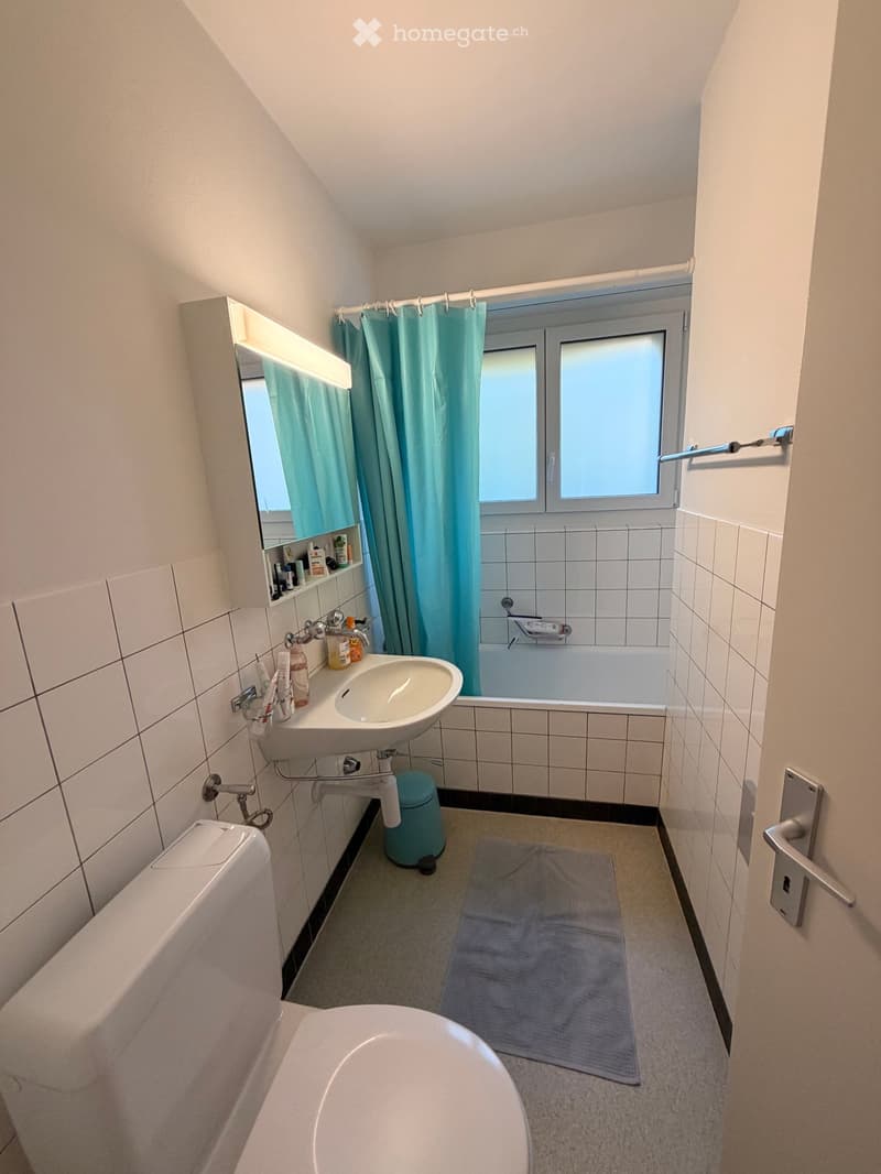 A compact bathroom with a sink, toilet, bathtub, and mirror. The space features tiled walls, a window with a blue shower curtain, and a small shelf holding toiletries.