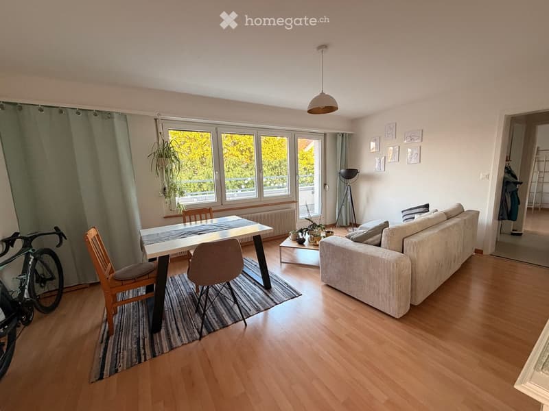 Bright living room with wooden flooring, large windows, a sofa, coffee table, dining table, and a bicycle near the wall.