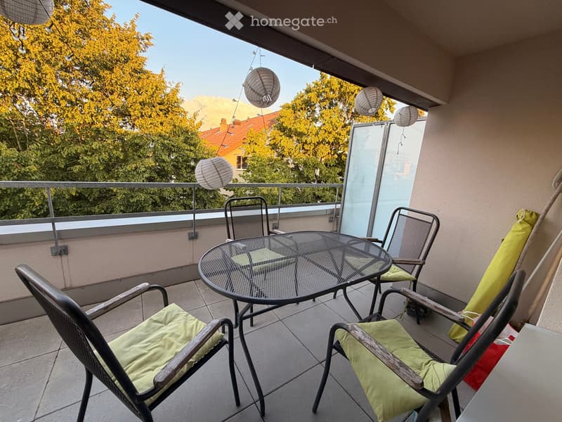 Balcony with a round glass table and four chairs, green cushions, hanging lanterns, and a view of trees and buildings.