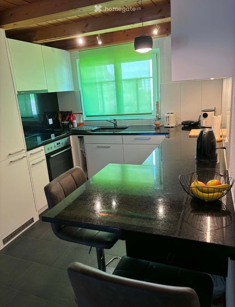 Modern kitchen with white cabinets, black countertops, and a central island with a black table and chairs. Appliances include an oven, sink, toaster, and blender. A fruit basket with bananas and apples is on the counter.