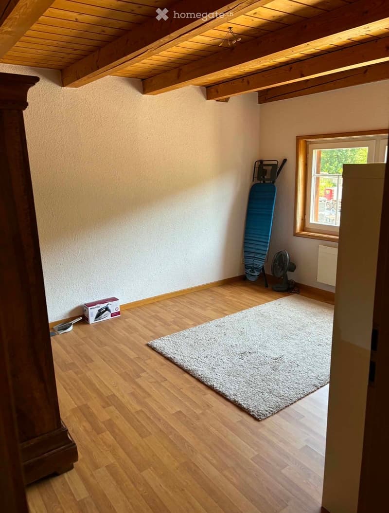 Empty room with wooden flooring, exposed wooden beams on the ceiling, a large window, and a small rug in the center.