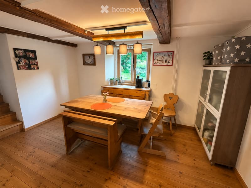 Cozy kitchen with wooden table, bench seating, chairs, and a cabinet. Exposed wooden beams on the ceiling and pendant lights hang above the table. A coffee machine and potted plants are on the counter.