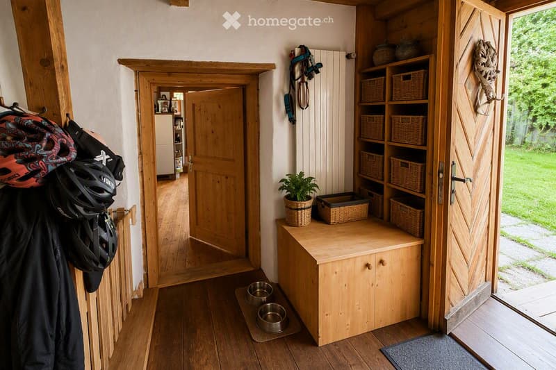 A wooden hallway with a built-in storage unit, helmets, jackets, and a small plant on the shelf. The hallway leads to another room and has a door to the outside.
