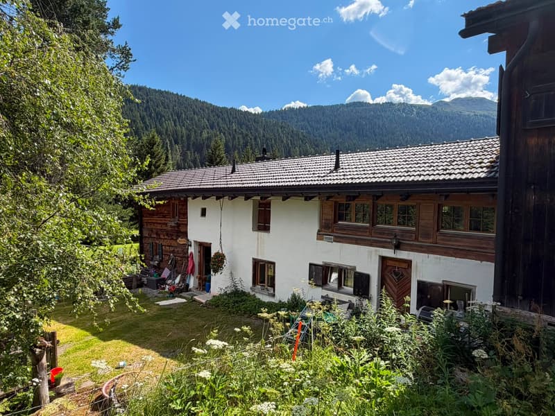 A two-story wooden house with white walls, surrounded by a lush garden and trees, with a scenic mountain backdrop.