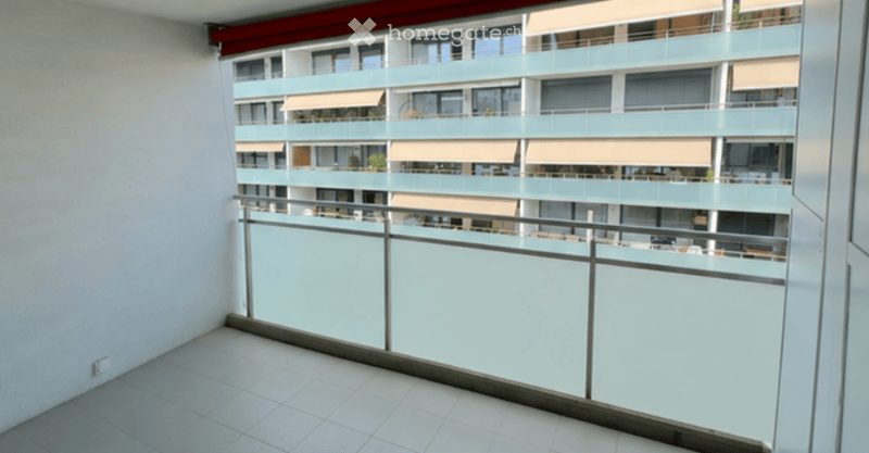 Balcony with modern glass railing, tiled floor, and view of a multi-story residential building with balconies and awnings.