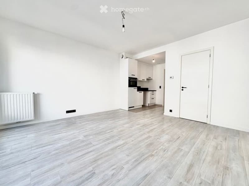 Empty room with white walls, light wooden flooring, and a white radiator on one wall. A doorway leads to a small kitchen area with white cabinets and a black oven.