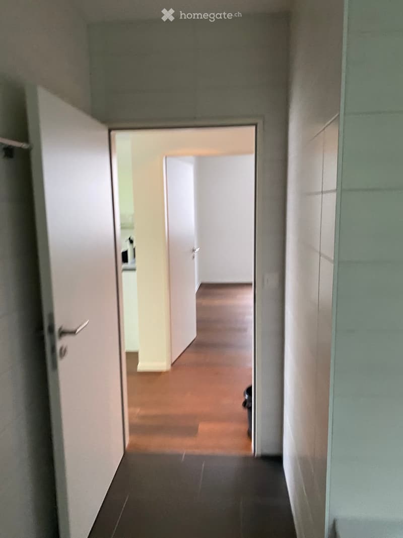 A narrow hallway with tiled flooring and white walls, featuring an open door leading to another room with wooden flooring visible in the background.