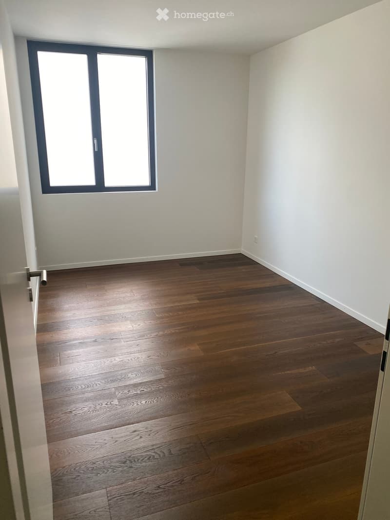 Empty room with wooden flooring and white walls, featuring a large window with double glass panels and a door handle visible on the left side.