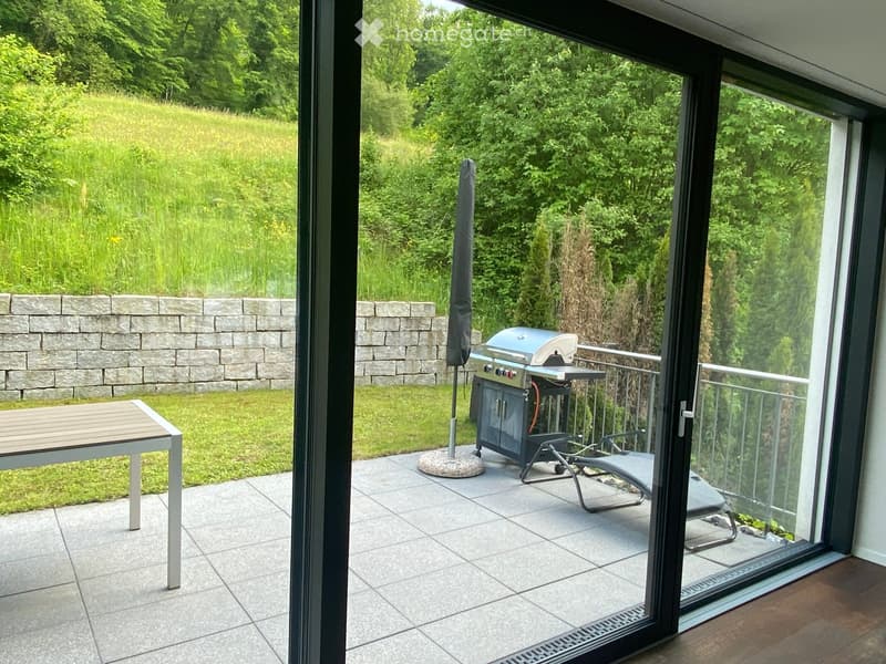 Balcony with tiled floor, outdoor table, chair, grill, and umbrella overlooking a green lawn and stone wall.