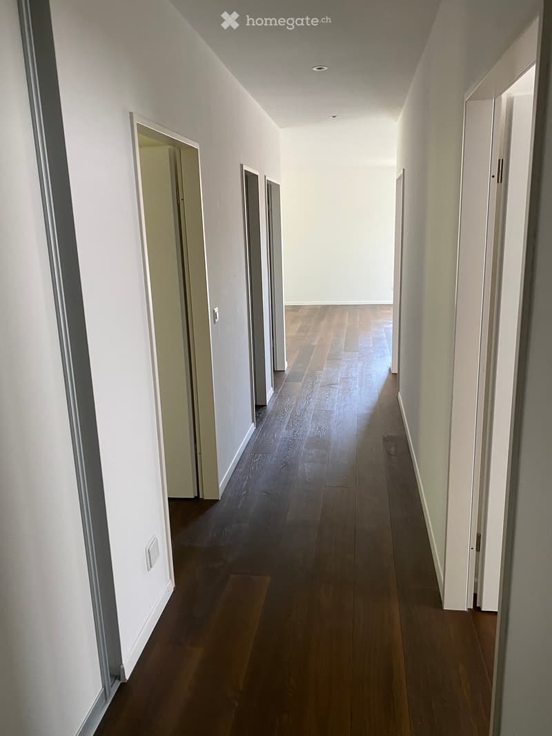 A long, narrow hallway with wooden flooring and white walls, featuring several doors on both sides and recessed ceiling lighting.