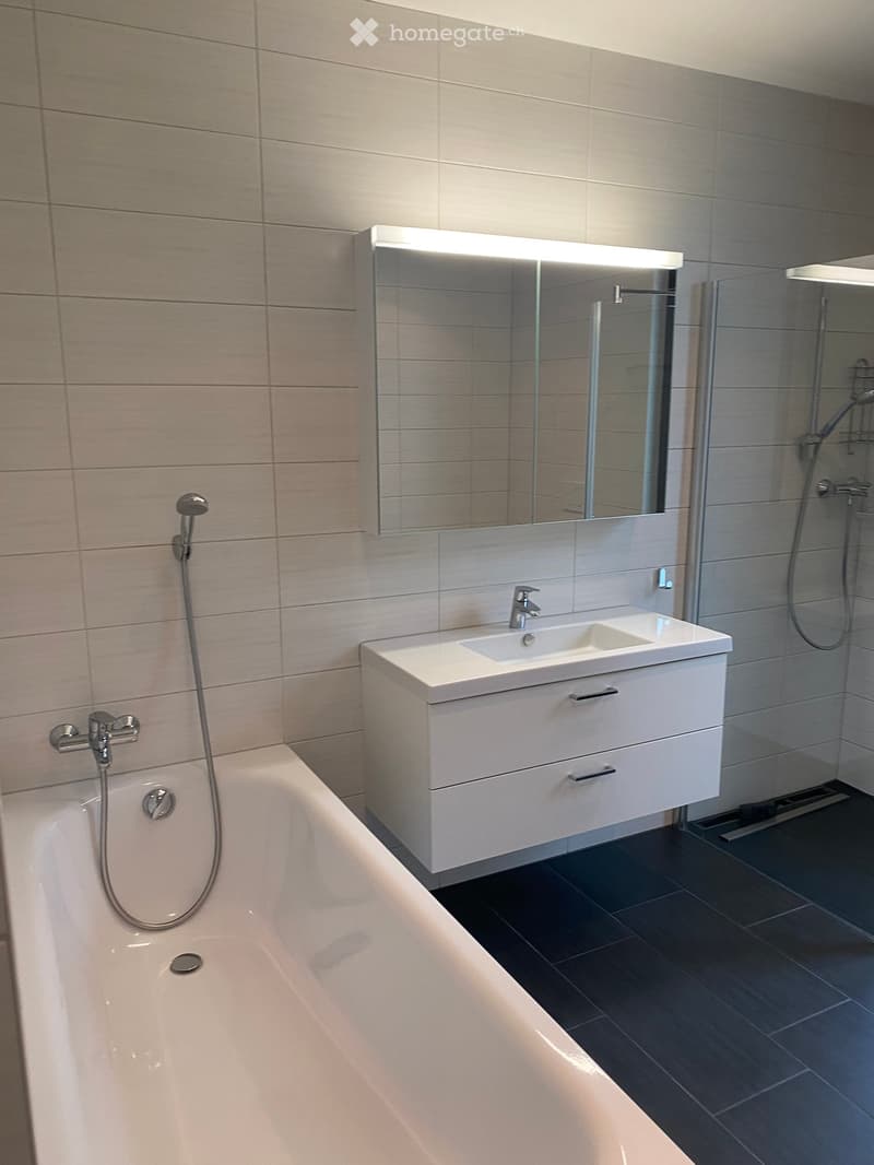 Modern bathroom with a bathtub, sink, and large mirror with built-in lighting. The walls are tiled in light gray, and the floor is dark gray tile.