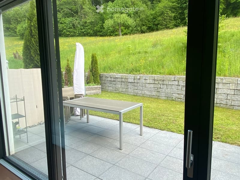 Balcony with tiled floor, outdoor table, chairs, and umbrella, overlooking a grassy garden with a stone wall.