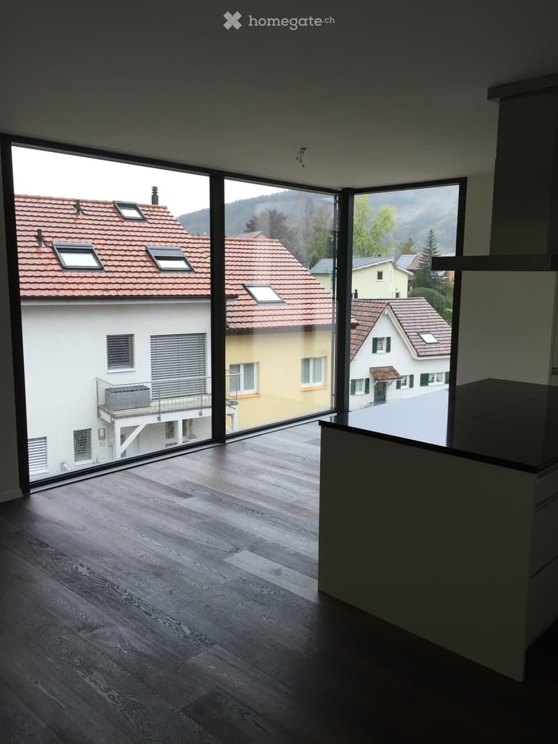 Empty room with wooden flooring and large glass windows offering a view of the surrounding houses and landscape.