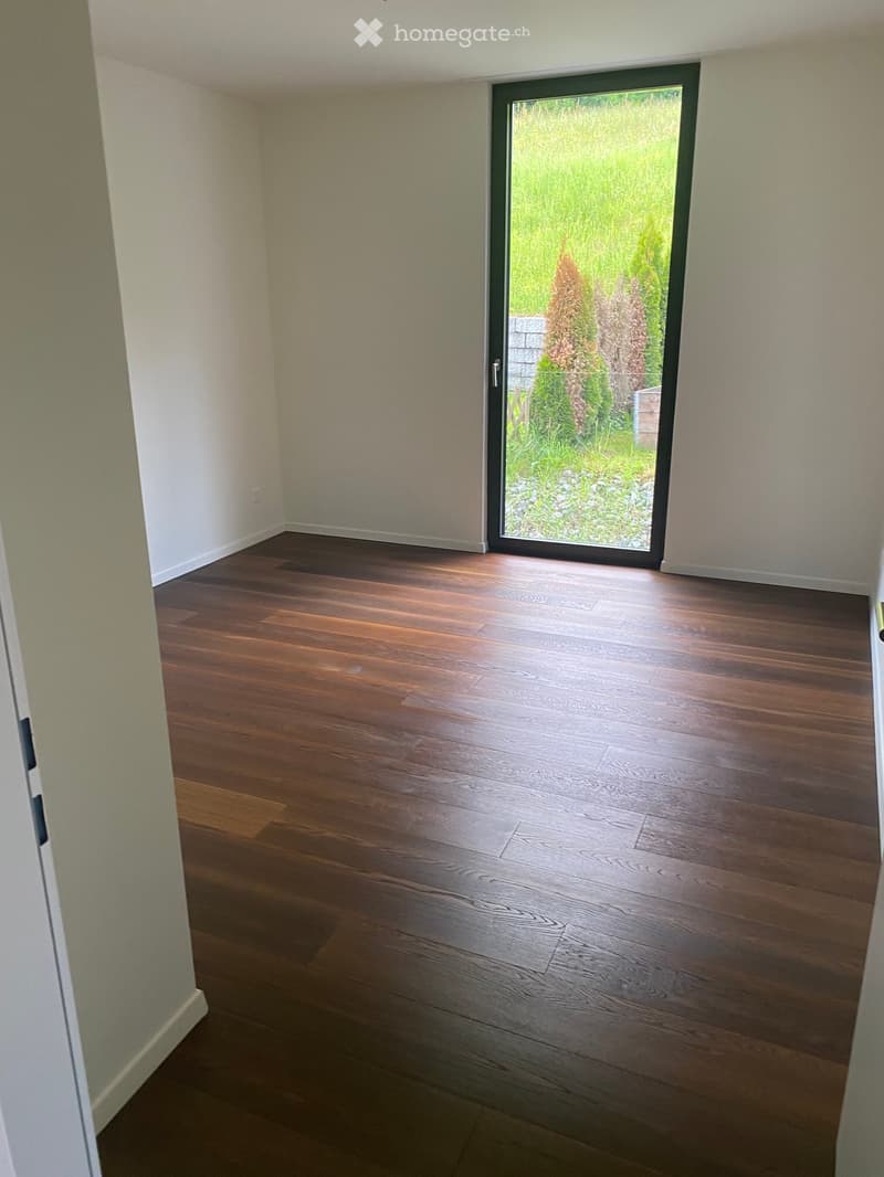 Empty room with wooden flooring, white walls, and a large sliding glass door leading to a green outdoor area.
