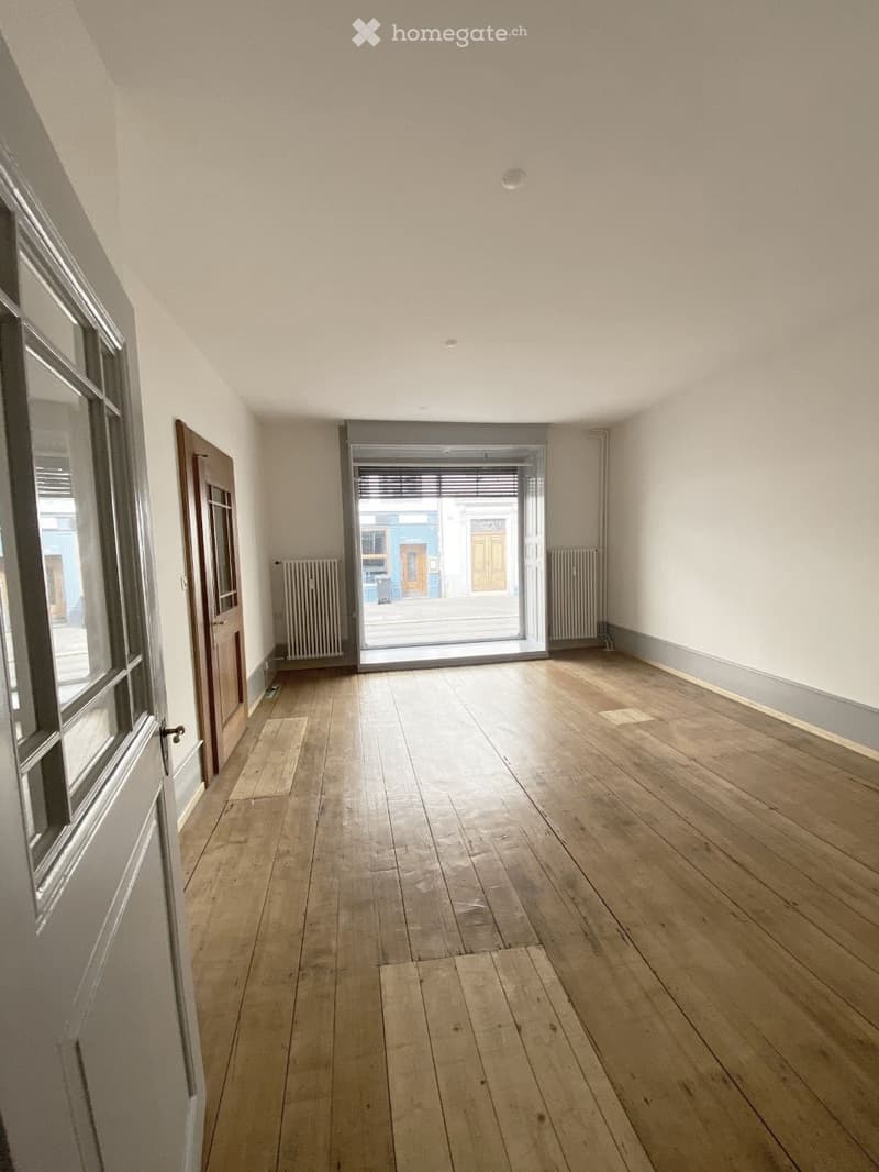 Empty room with wooden flooring, white walls, and a large window with blinds. There are two doors, one leading to another room and the other to an outdoor area.