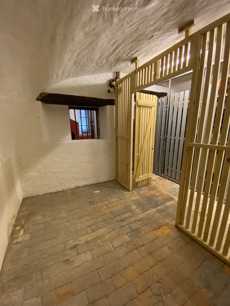 A narrow corridor with exposed stone walls and a brick floor, featuring a wooden gate and a small window with stained glass.