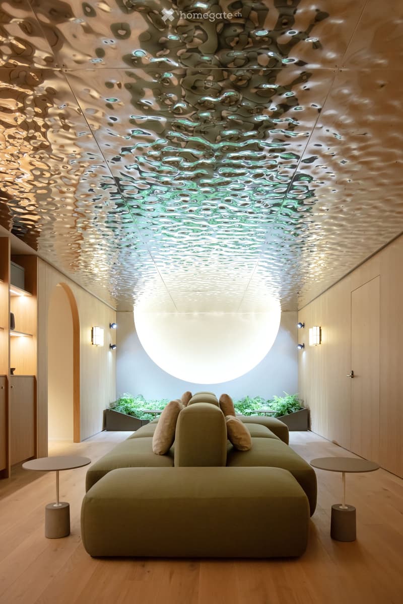 A modern living room with a large green sofa, two side tables, a wooden floor, and a unique ceiling design. There is a small garden area with plants visible through a window.