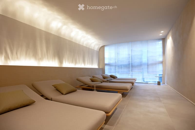 A room with several lounge chairs, beige walls, and a large window with curtains.