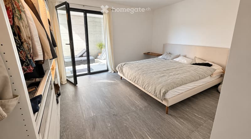 Modern bedroom with a large bed, nightstand, and a sliding glass door leading to a balcony area with outdoor furniture.
