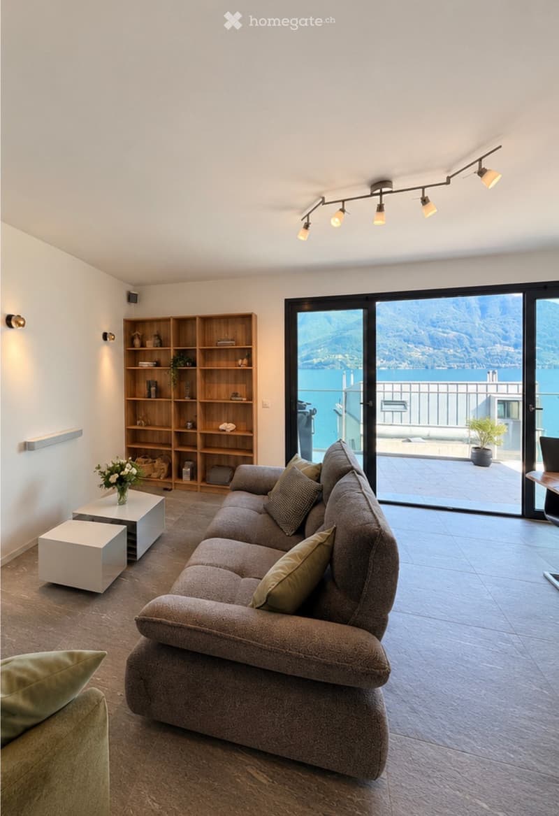 Modern living room with a grey sectional sofa, coffee table, bookshelf, and a vase with flowers. Large sliding glass doors open to a balcony with a scenic mountain view.