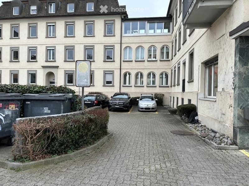 Multi-story residential building with multiple windows and a parking area in front.