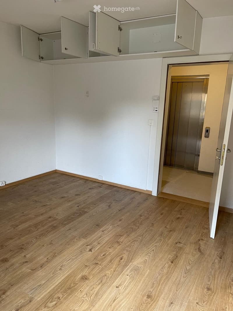 Empty room with wooden flooring, white walls, and an open door leading to an elevator area.