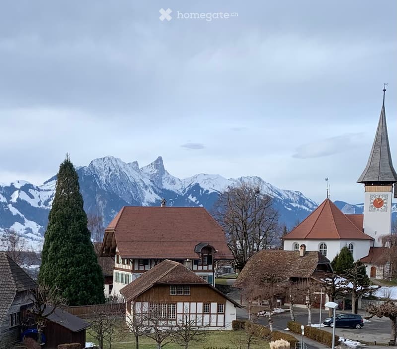 Village scene with traditional houses, a church tower, and snow-capped mountains in the background.