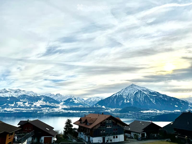 A cluster of traditional Swiss houses with red-tiled roofs and white walls, situated on a hillside overlooking a lake and snow-capped mountains.