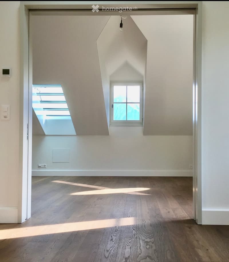 Empty room with wooden flooring, white walls, and large windows allowing natural light to enter. The room features a modern design with a visible electrical outlet and thermostat on the wall.